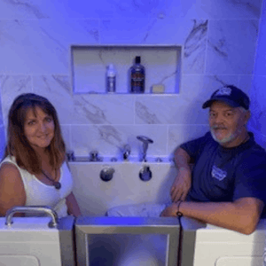 Couple smiling inside an Ella walk-in tub with blue ambient lighting and marble tile wall with built-in shelf