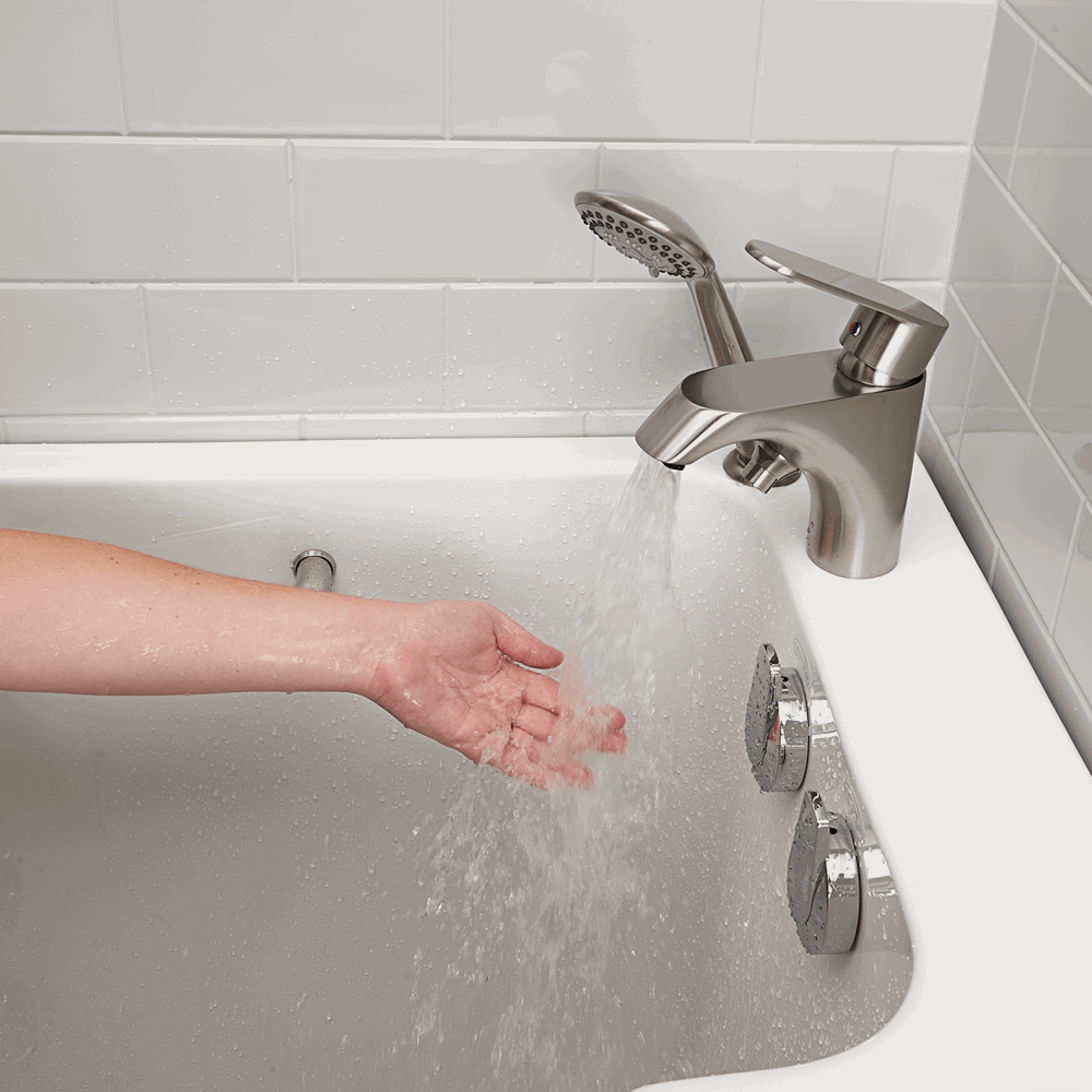 Mano comprobando el agua de un grifo de níquel cepillado dentro de una bañera con puerta Ella Lounger blanca con teleducha y mandos de los chorros en cromado
