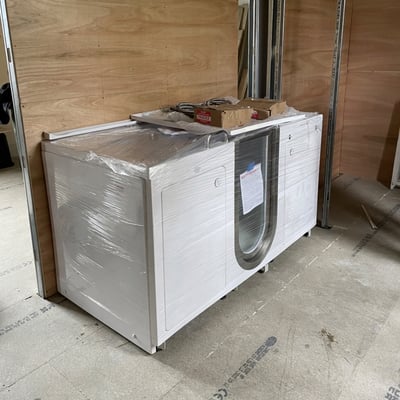 Baignoire à porte Ella emballée dans un film plastique de protection avec accessoires posés dessus, prête pour la livraison et l'installation