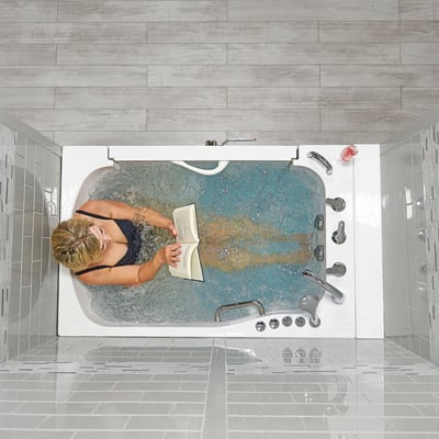 Senior woman reading a book while soaking in an Ella TransferXXL walk-in tub with active hydrotherapy jets in a modern gray-tiled bathroom