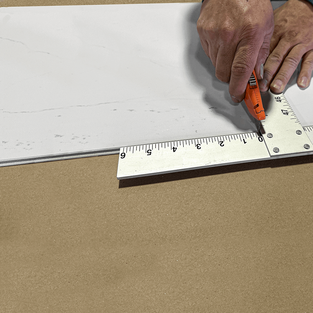 Installer scoring a Carrara marble look grout-free shower wall tile with a utility knife along a straight edge