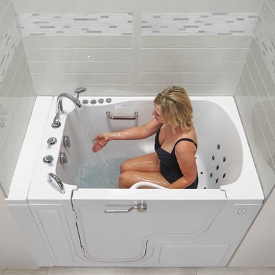 Woman seated in a white walk-in tub with outward swing door, water jets, and chrome faucet running water