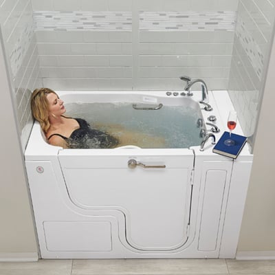 Woman relaxing in a white walk-in tub with outward swing door, hydrotherapy jets, chrome faucet, and a book and wine glass on