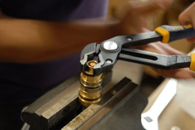 Technician using pliers to tighten a brass thermostatic control valve cartridge secured in a metal vise during walk-in tub assembly