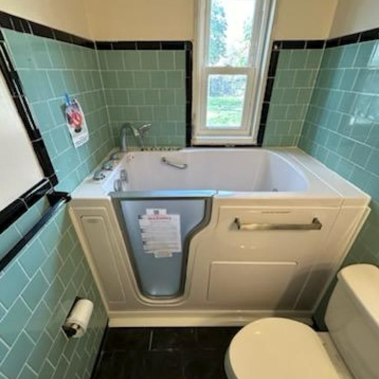 Ella walk-in tub installed in a teal-tiled bathroom with side entry door, grab bar, and faucet controls near a window