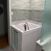 White Ella walk-in tub installed in a modern bathroom with pebble mosaic tile accent wall and glass panel divider