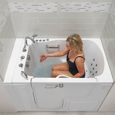 Mujer con bañador negro probando el flujo de agua caliente del grifo sentada en una bañera con puerta Ella Transfer XL con jets de hidroterapia