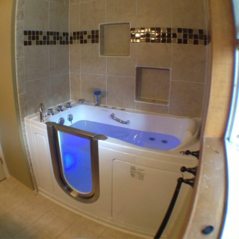 Walk-in tub with glowing blue chromotherapy lighting installed in a tiled bathroom with built-in wall niches and jets visible