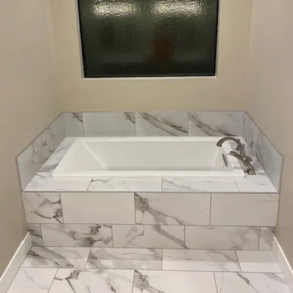 White soaking tub with bronze faucet set into a marble-tile surround with matching floor and frosted window above