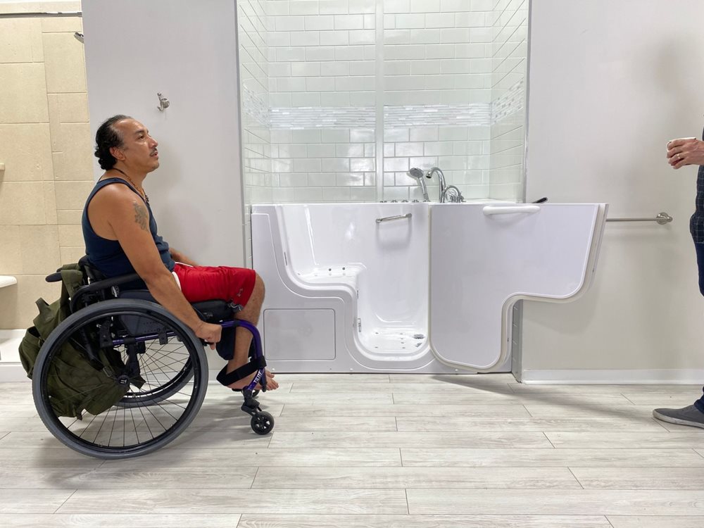 Utilisateur en fauteuil roulant examinant une baignoire à porte Ella, porte latérale ouverte et barres d'appui, lors d'une consultation en showroom