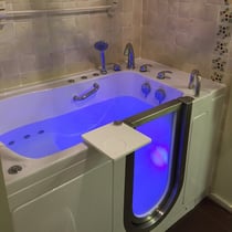 Ella walk-in tub with blue chromotherapy lighting, open door, swivel tray, jets, and chrome faucet set in tiled bathroom