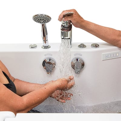 Senior bathing hands under running water from chrome faucet controls on an Ella walk-in tub with handheld showerhead