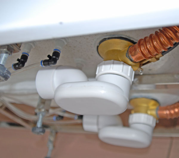 Primer plano del codo de desagüe de PVC blanco y de las conexiones de fontanería debajo de una bañera con puerta Ella de modelo antiguo