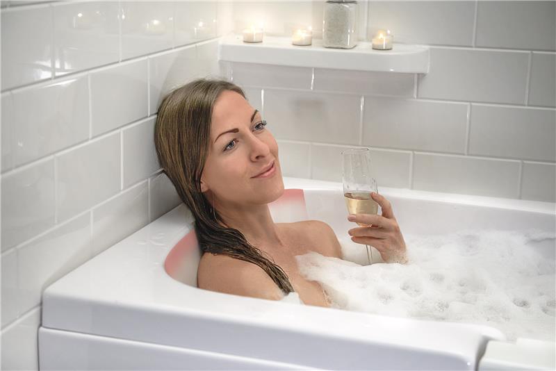Une femme se détend dans une baignoire à porte avec des jets de bulles, tenant un verre de vin, avec des bougies allumées sur le rebord de la baignoire