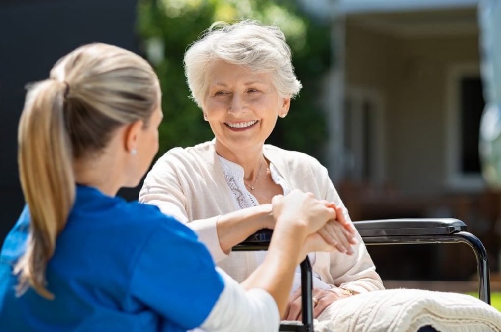 Smiling senior woman in wheelchair holding hands with a caregiver in blue scrubs outdoors at an assisted living facility