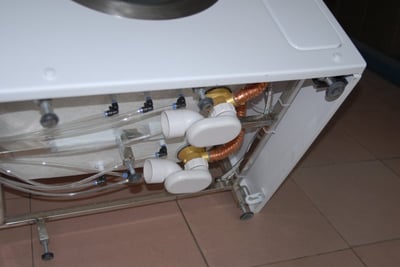 Underside view of an Ella walk-in tub showing internal plumbing with copper coils, white PVC valves, and clear drain tubing components