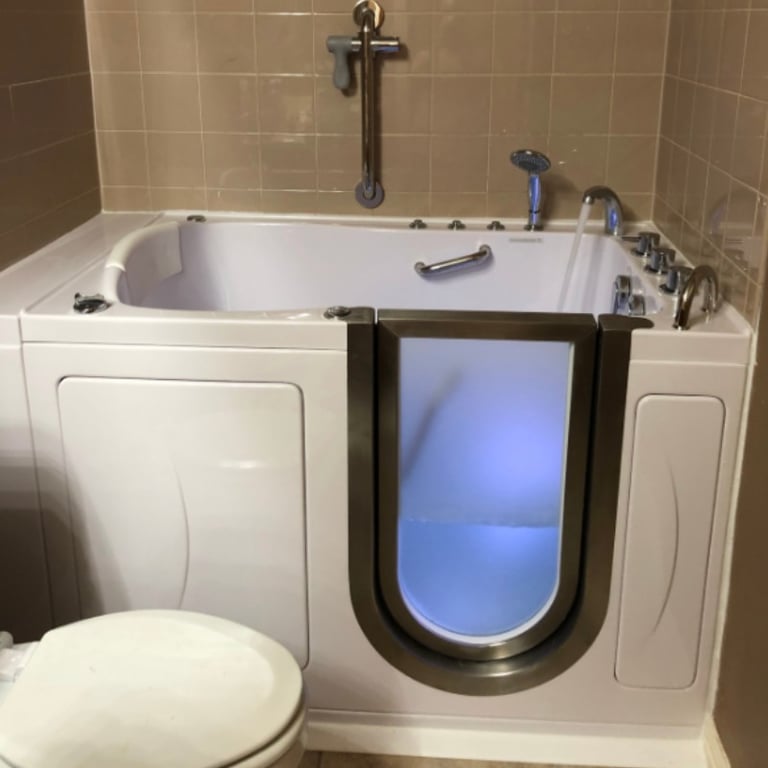 Ella walk-in tub with open outward swing door showing blue interior lighting, grab bars, and chrome faucet fixtures installed in a beige tiled bathroom