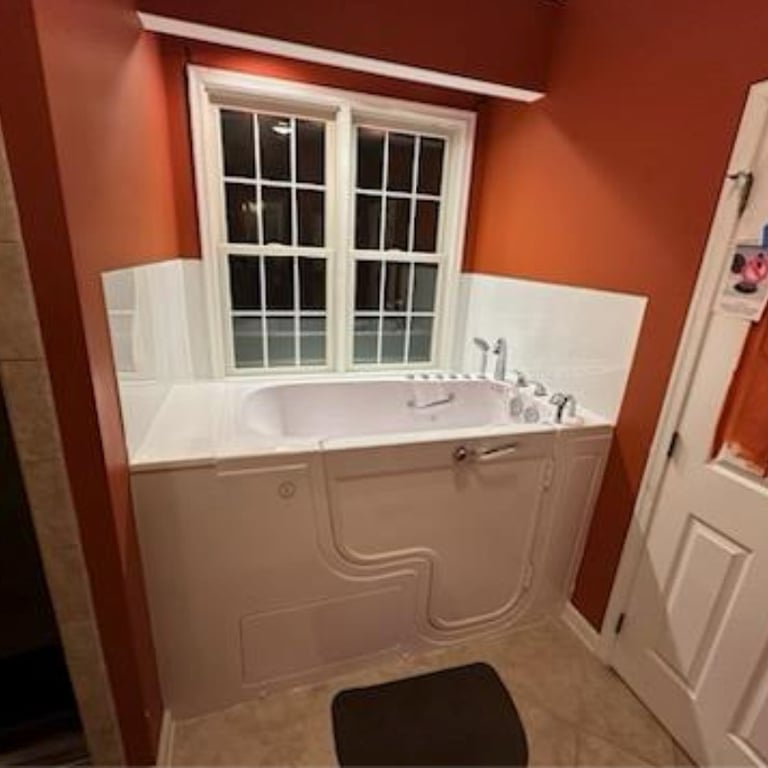 Ella walk-in tub installed in a rust-red bathroom with white tile surround, chrome faucets, and a safety grab bar on the door