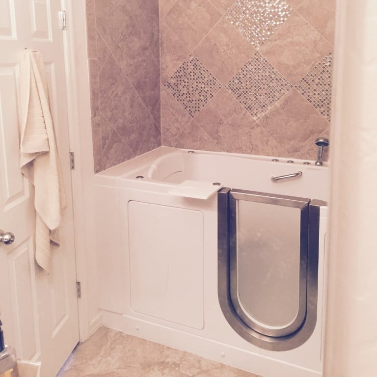 White Ella walk-in tub with stainless steel door installed against beige diamond-pattern tile walls in a home bathroom
