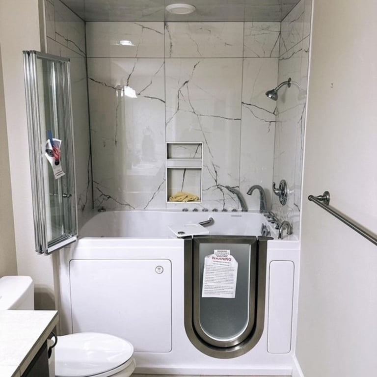 Ella walk-in tub installed in a bathroom with marble-look surround walls, shower head, built-in niche, and folding glass door panel