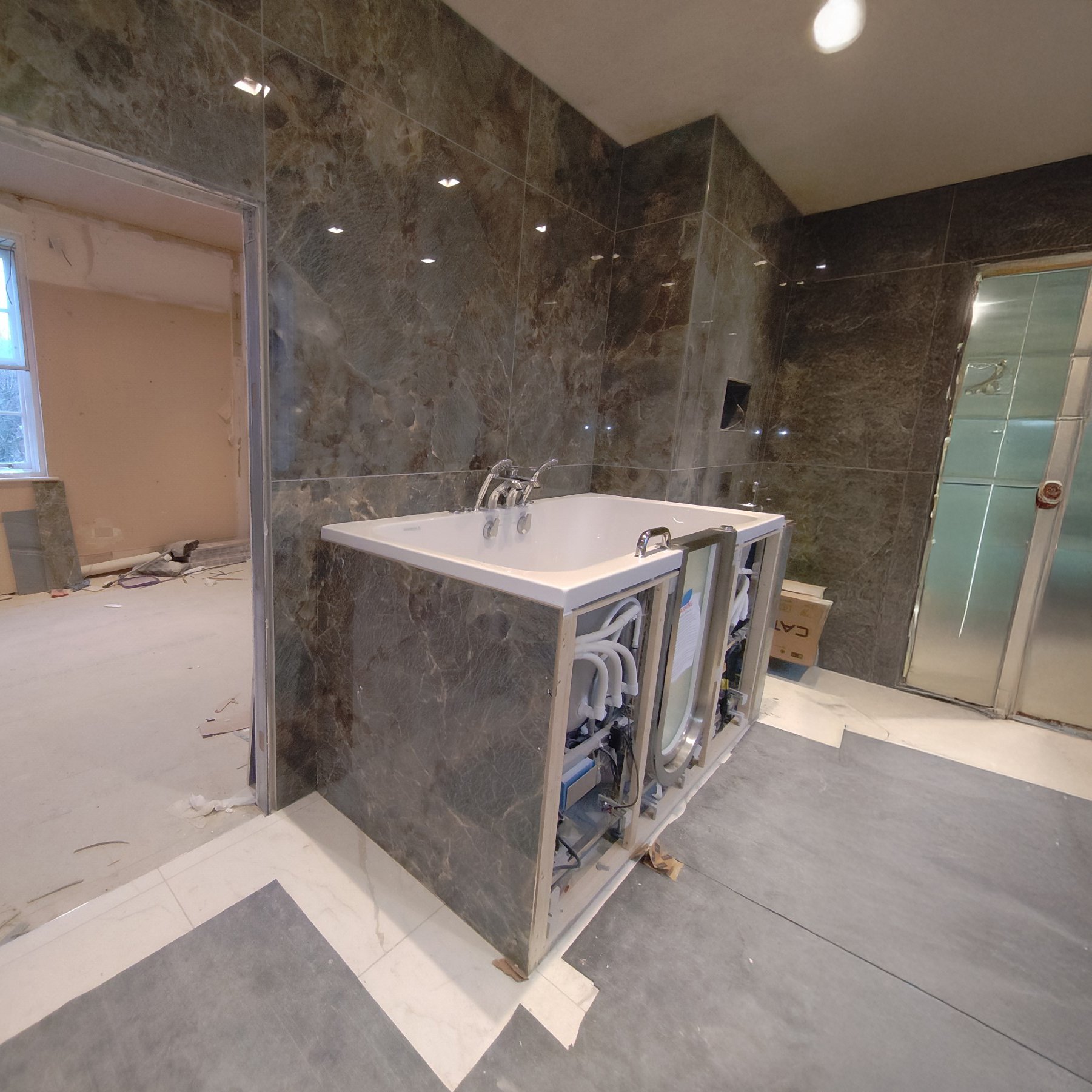 Baignoire à porte Ella en cours d'installation, panneaux déposés, laissant voir le châssis, la plomberie et les jets devant des murs carrelés