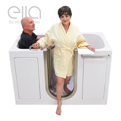 Senior woman in yellow robe stepping into the Ella Companion walk-in tub while a man soaks inside the dual-entry bathing area