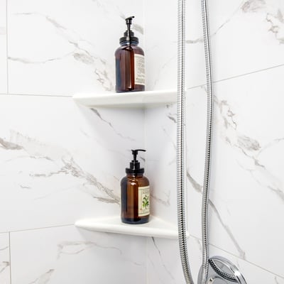 Two white built-in corner shelves holding amber soap bottles inside an Ella accessible shower stall with marble-look walls