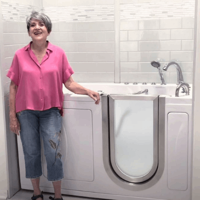 Mujer mayor con blusa rosa sonriendo junto a una bañera con puerta de Ella con puerta de apertura hacia fuera en un baño blanco alicatado