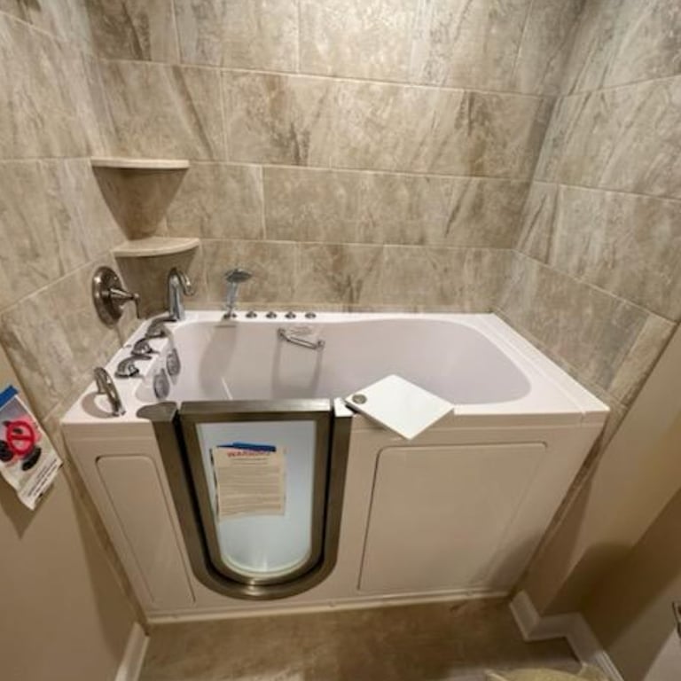 Ella walk-in tub with stainless steel door installed in a tiled bathroom alcove with chrome faucets and corner shelves