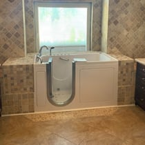 White Ella walk-in tub installed in a tiled bathroom alcove with stone mosaic surround and a window backdrop