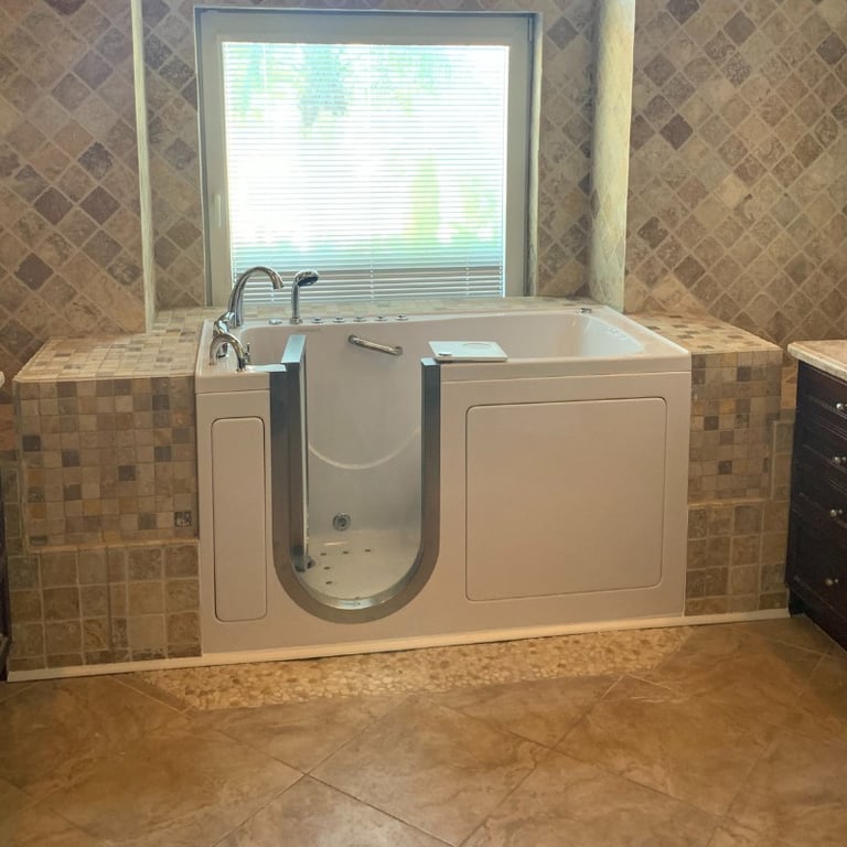 White Ella walk-in tub installed in a tiled bathroom alcove with stone mosaic surround and a window backdrop