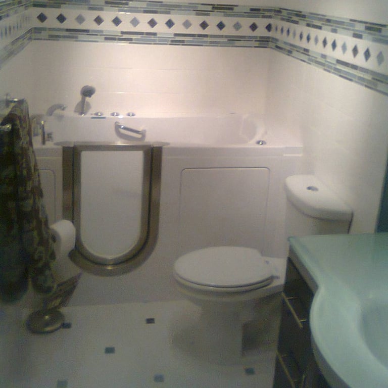 Walk-in tub with gold grab bar door handle installed in a tiled accessible bathroom with toilet and decorative mosaic border