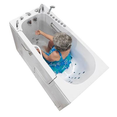 Senior woman in blue floral top seated inside the Ella Transfer60 wheelchair accessible walk-in bathtub adjusting faucet controls from open outward swing door side
