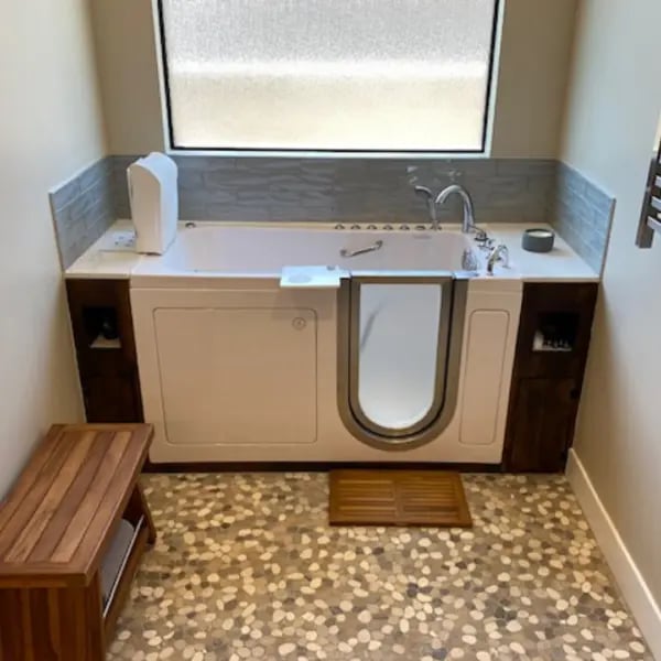 White walk-in tub installed in a bathroom with pebble tile floor, wood storage cabinets, and a teak bench nearby