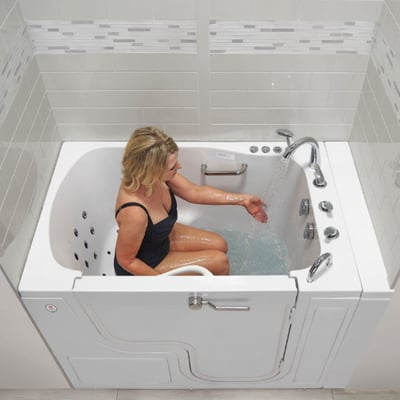 Woman seated inside a white walk-in tub with outward swing door, reaching toward running water from a chrome faucet with jets