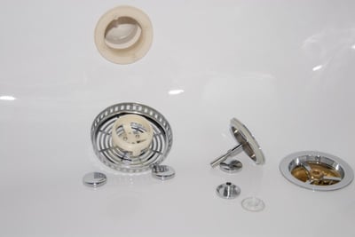 Close-up of walk-in tub drain cover components including chrome strainer basket, stopper, and plastic collar on white tub floor