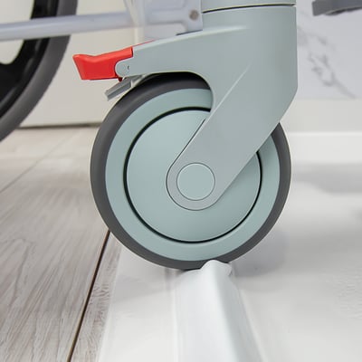 Close-up of a locking caster wheel with red brake lever on a roll-in shower chair at the accessible shower entry threshold