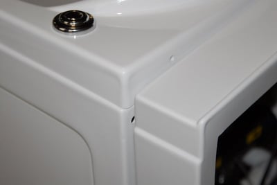 Close-up of white walk-in tub extension panel kit corner showing glossy acrylic finish and chrome drain fitting detail