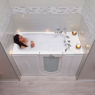 Woman relaxing with a drink in a bubble-filled Ella Capri walk-in bathtub surrounded by candles and white tile walls
