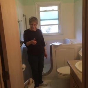 Senior woman named Norma standing in her remodeled bathroom beside a newly installed walk-in tub with open door
