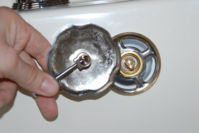 Hand lifting a chrome Ella walk-in tub drain cover to reveal the brass drain opener mechanism beneath
