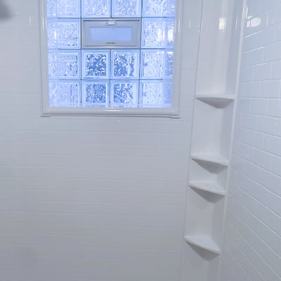 White accessible shower stall interior with built-in corner shelves and frosted glass block window providing natural light