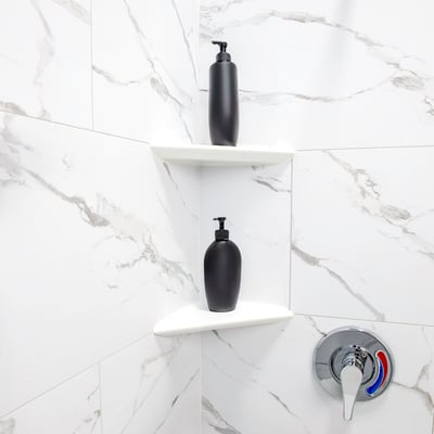 Two white acrylic corner shelves holding black soap dispensers inside a marble-look accessible shower stall with chrome controls