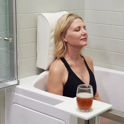 Woman relaxing with eyes closed in an Ella walk-in tub featuring a padded removable headrest and neck support pillow with a drink on the tray