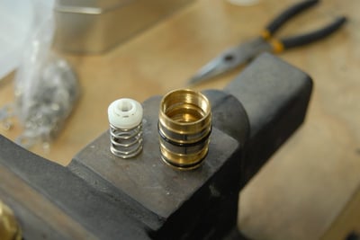 Disassembled brass thermostatic control valve cartridge with spring and ceramic seat resting on a workshop vise