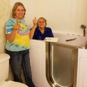 Two happy women giving thumbs up next to an Ella's Bubbles walk-in tub with stainless steel door and grab bar