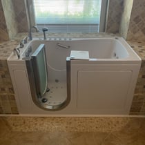 White Ella walk-in tub with open door showing air jets, grab bar, swivel tray, and chrome faucet installed in tiled bathroom