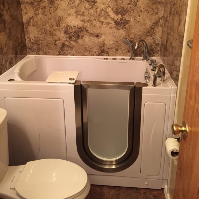 White Ella walk-in tub with outward swing door installed in a bathroom with stone wall surround and chrome faucet fixtures