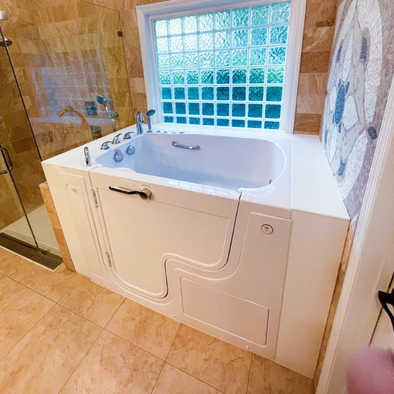 White Ella walk-in tub with inward swing door and chrome grab bar installed in a tan tile bathroom with glass block window