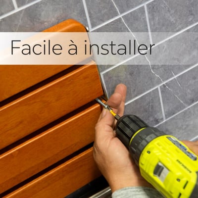 Installateur utilisant une perceuse électrique pour monter un siège de douche pliant en teck sur un mur en carrelage gris, avec le texte « Easy to Install »